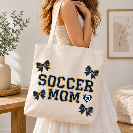 Person holding a tote bag with 'Soccer Mom' text and blue bows in a home setting.