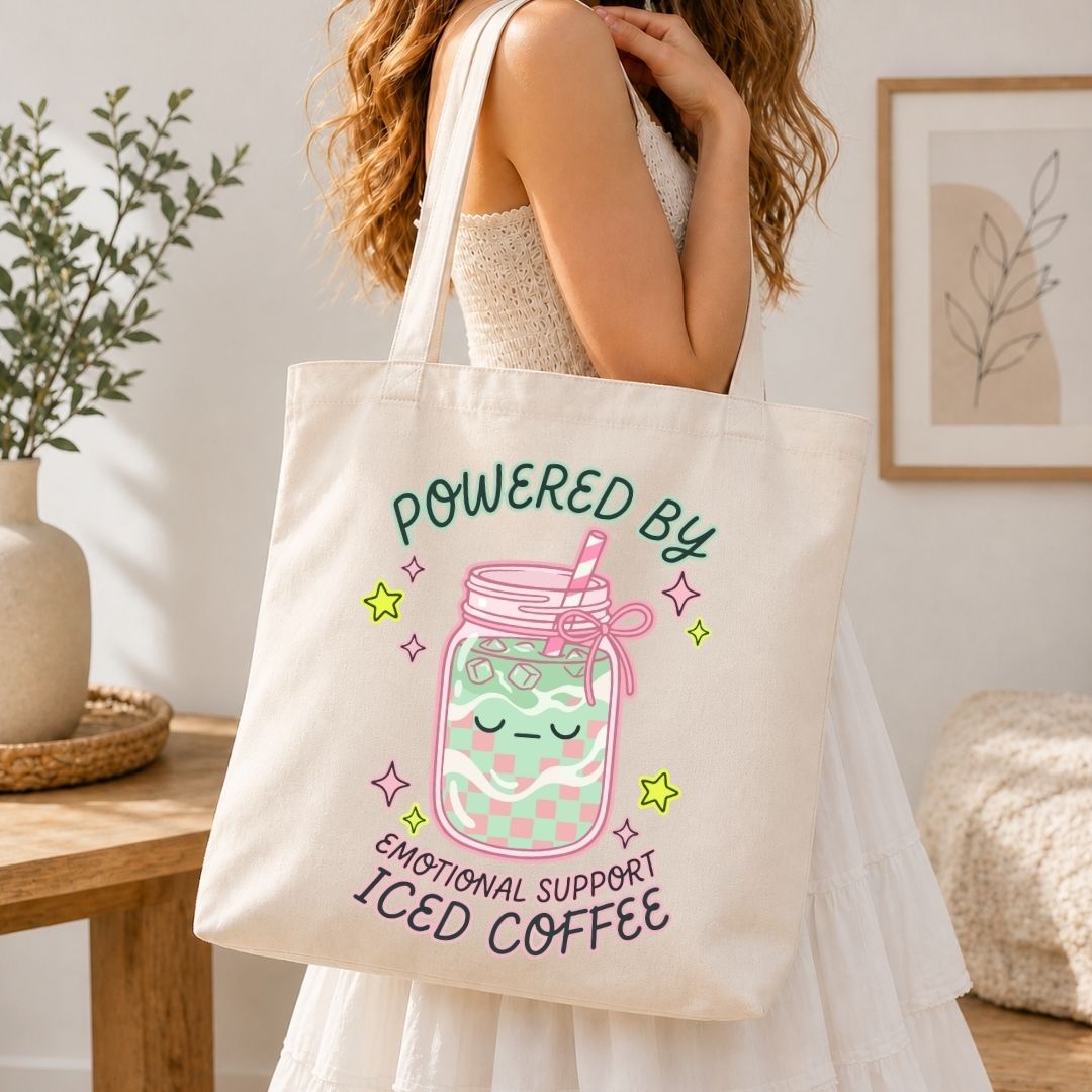 Person holding a tote bag with a design of a jar of iced coffee and text 'Powered by Emotional Support Iced Coffee' in a home setting.