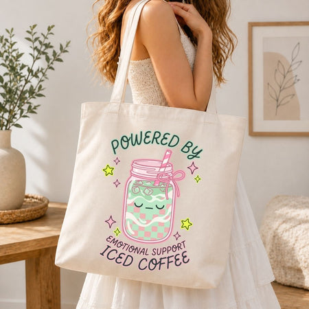 Person holding a tote bag with a design of a jar of iced coffee and text 'Powered by Emotional Support Iced Coffee' in a home setting.