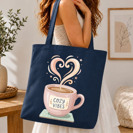 Navy tote bag with a pink coffee cup design and 'Cozy Vibes' text, held by a person in a home setting.
