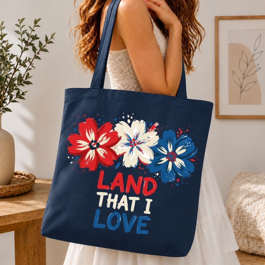 Navy tote bag with floral design and 'Land That I Love' text held by a person in a home setting.