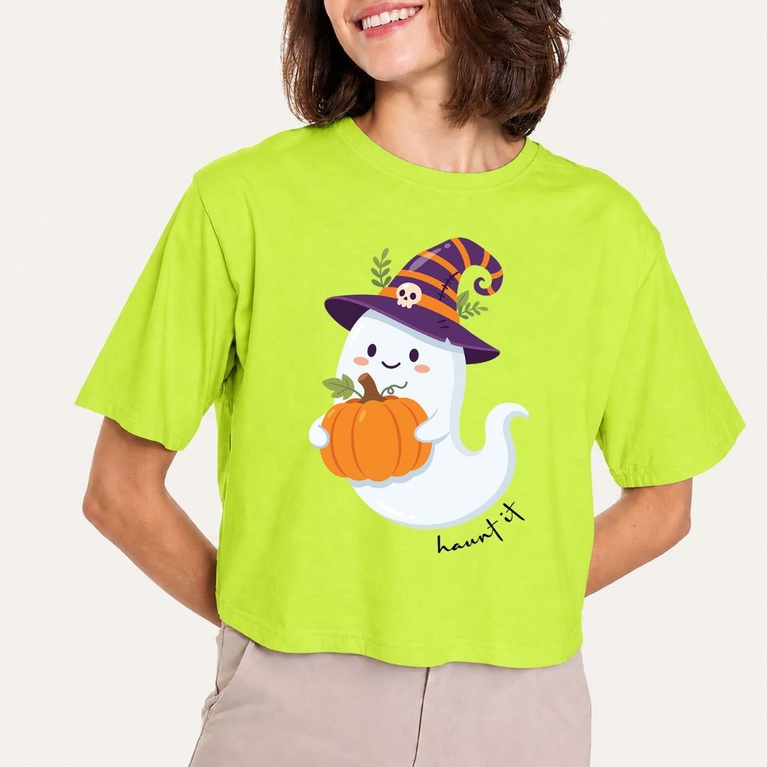 A woman wearing a bright lime green cropped t-shirt with a cartoon graphic of a white ghost holding an orange pumpkin and wearing a purple witch hat. The shirt has the word 'haunted' printed in black script below the graphic.