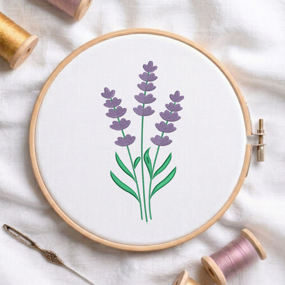 A wooden embroidery hoop holding white fabric with a stitched lavender flower design in purple and green, surrounded by spools of thread and a needle on a white fabric background.