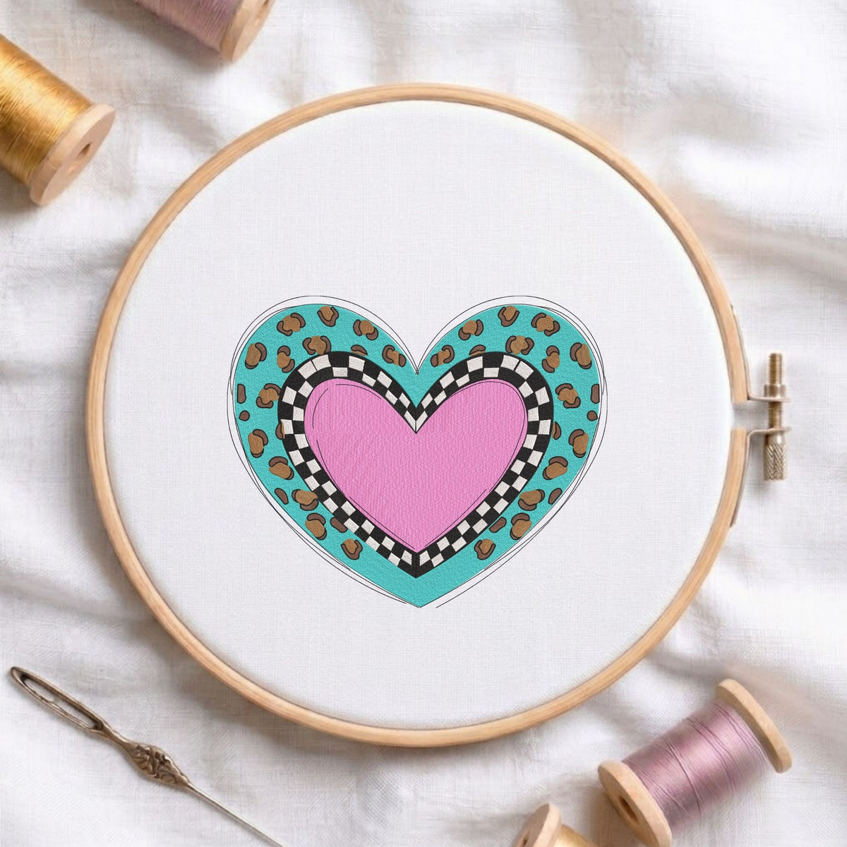  Layered pink heart with a black and white checkerboard pattern and turquoise leopard print machine embroidery design displayed in a wooden embroidery hoop on white fabric with thread spools and a needle   