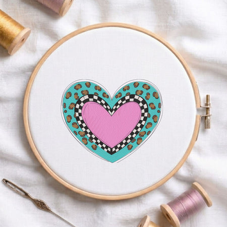  Layered pink heart with a black and white checkerboard pattern and turquoise leopard print machine embroidery design displayed in a wooden embroidery hoop on white fabric with thread spools and a needle   