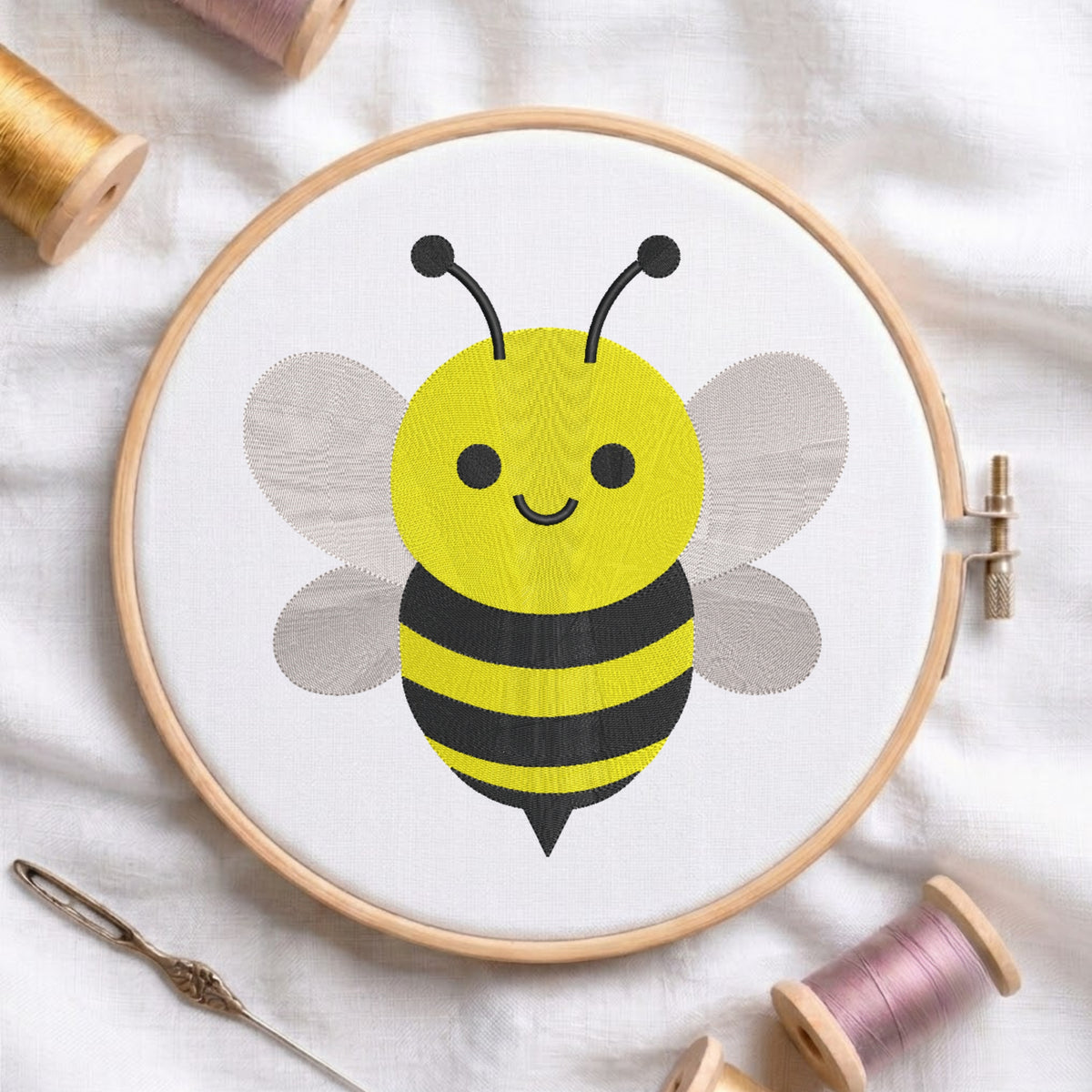 A wooden embroidery hoop holds white fabric with a cartoon bumblebee design in yellow, black, and gray. The hoop is photographed on a white fabric surface with thread spools and a needle around it.