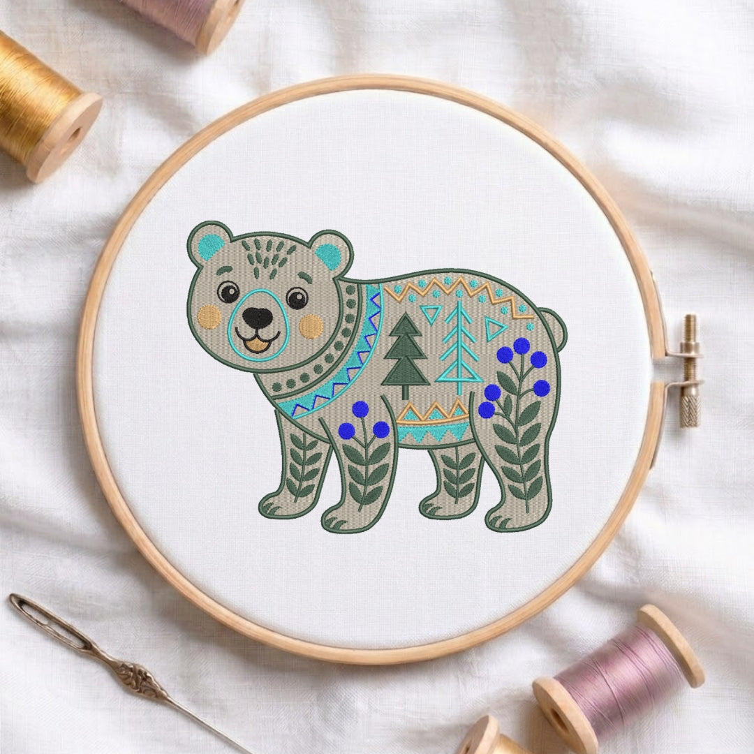 A circular embroidery hoop holding white fabric with a stylized embroidered bear design. The bear has a gray body with teal, beige, and green geometric patterns, blue berries, and green pine trees.