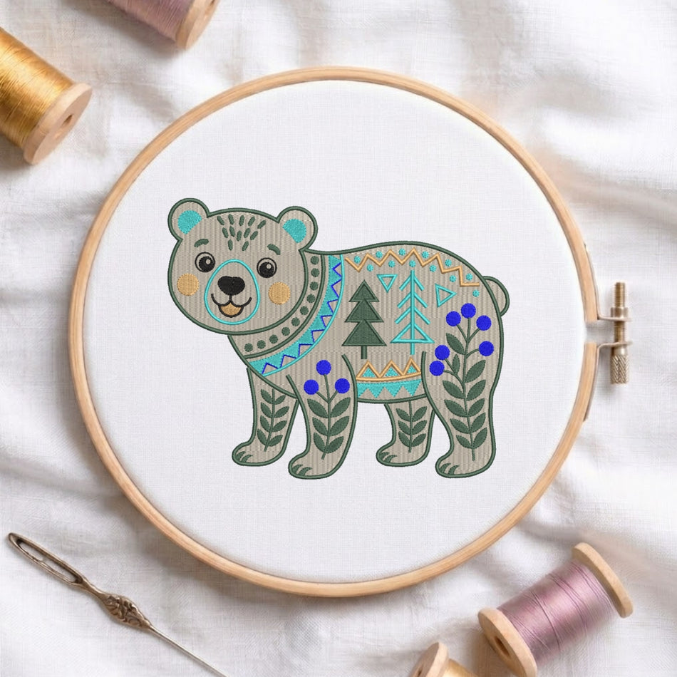 A circular embroidery hoop holding white fabric with a stylized embroidered bear design. The bear has a gray body with teal, beige, and green geometric patterns, blue berries, and green pine trees.