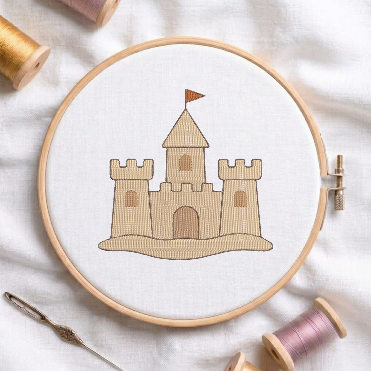 A wooden embroidery hoop holding white fabric with a stitched tan castle design and small orange flag, surrounded by spools of thread and a needle on a white fabric background.