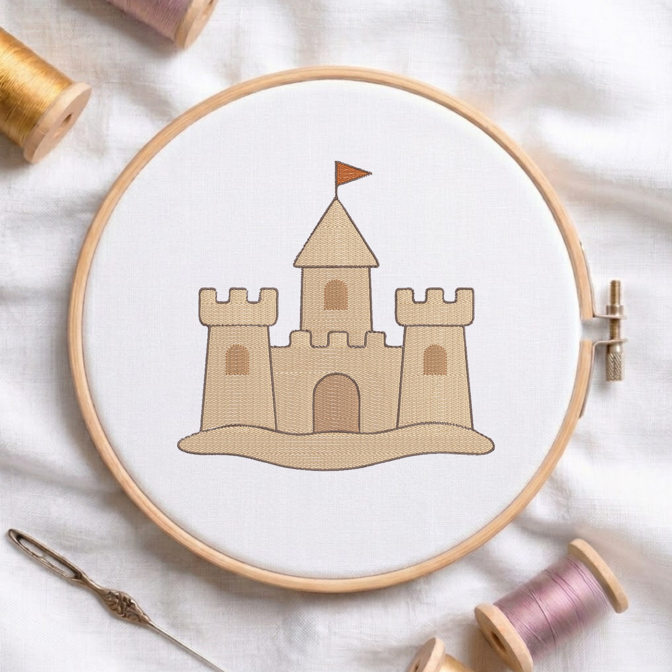 A wooden embroidery hoop holding white fabric with a stitched tan castle design and small orange flag, surrounded by spools of thread and a needle on a white fabric background.