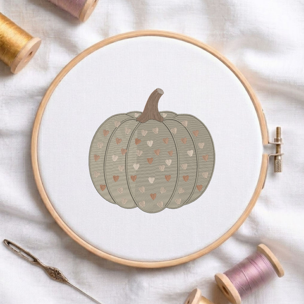 A circular embroidery hoop holding white fabric with a stitched pumpkin design in muted gray-green, brown, and small peach hearts. Spools of thread and a needle are arranged around it on a white fabric background.