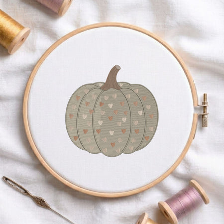A circular embroidery hoop holding white fabric with a stitched pumpkin design in muted gray-green, brown, and small peach hearts. Spools of thread and a needle are arranged around it on a white fabric background.