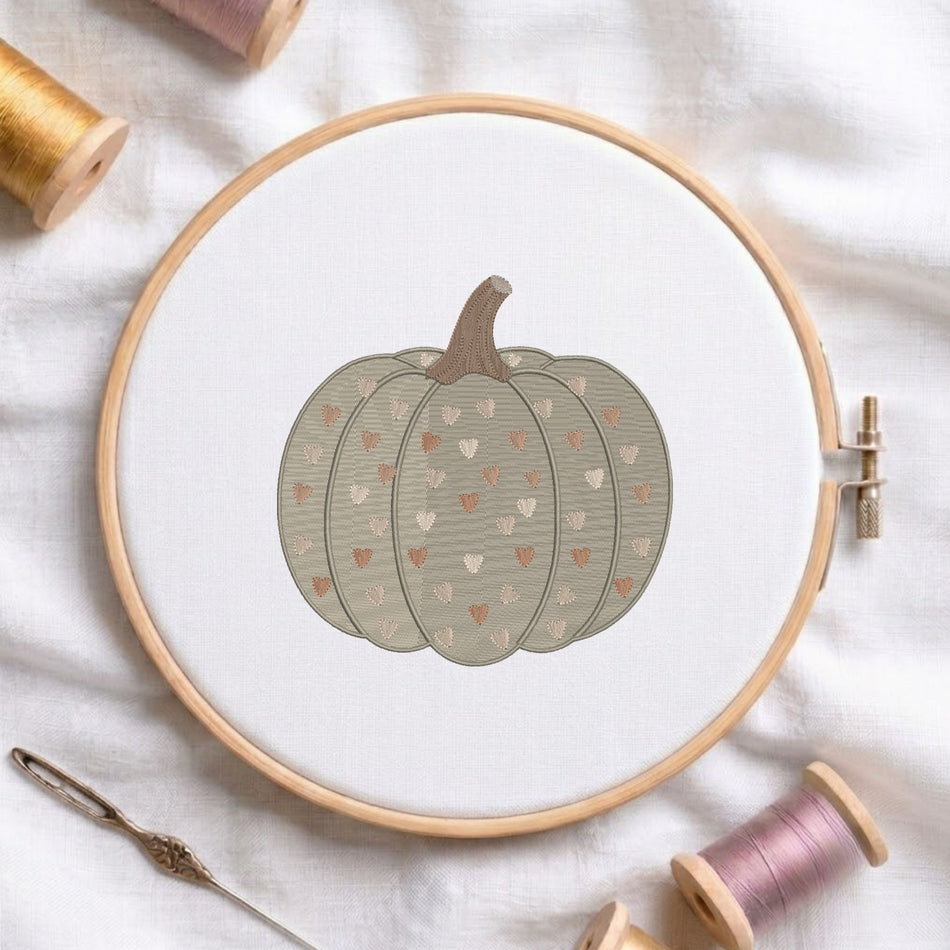 A circular embroidery hoop holding white fabric with a stitched pumpkin design in muted gray-green, brown, and small peach hearts. Spools of thread and a needle are arranged around it on a white fabric background.