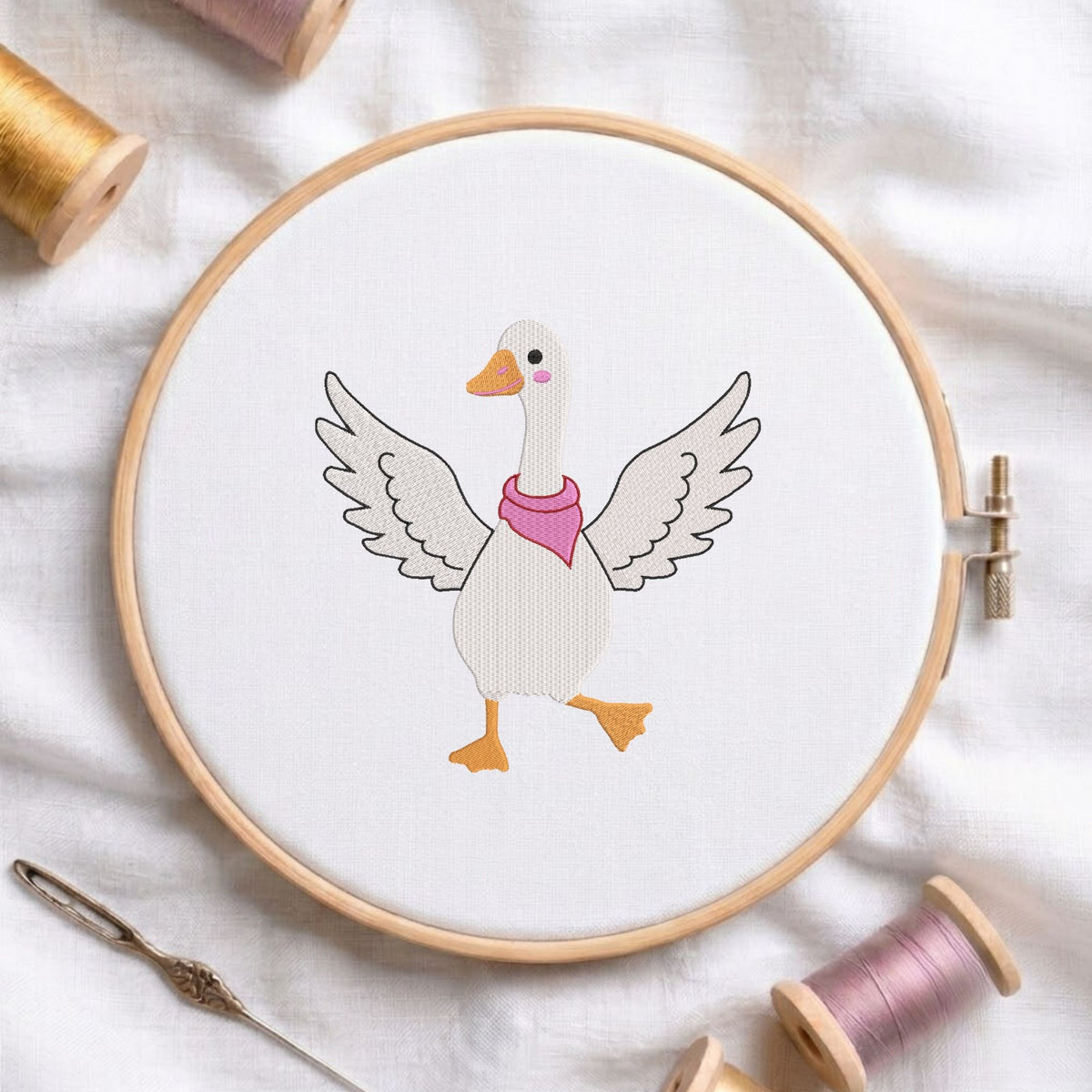 Cute dancing goose with open wings and a pink scarf machine embroidery design displayed in a wooden hoop on white fabric with thread spools and a needle