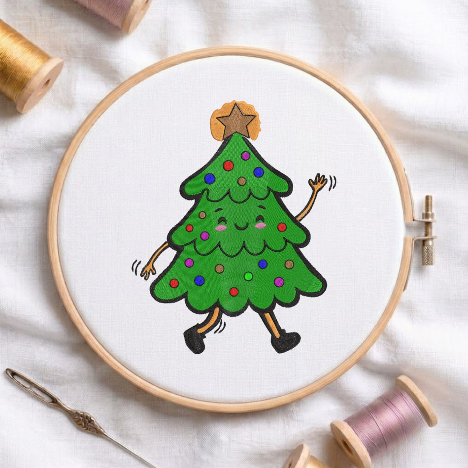 A circular embroidery hoop holding white fabric with a cartoon Christmas tree design in green, black, orange, purple, and red. The hoop is photographed on a white fabric surface with spools of thread and a needle around it.