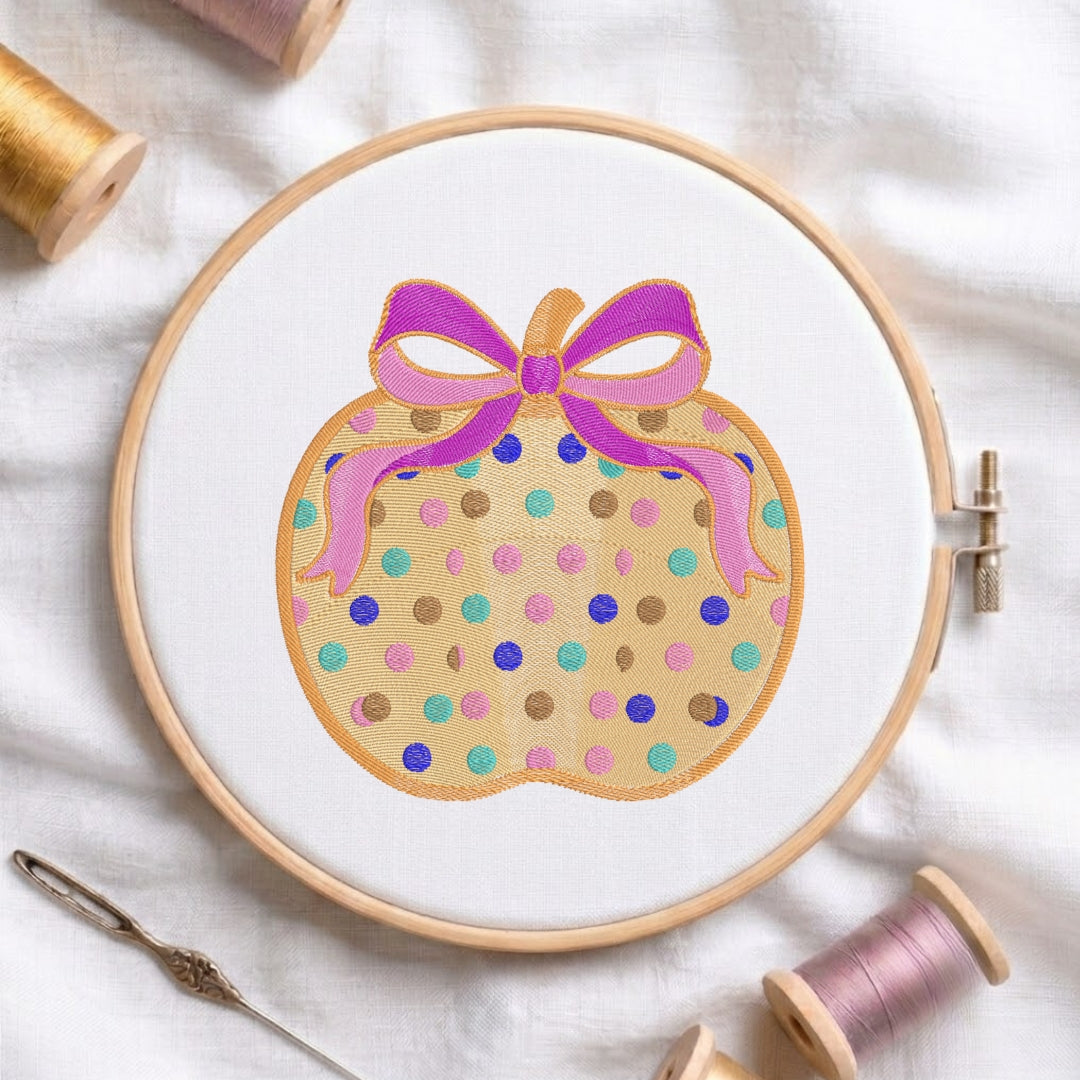 A circular embroidery hoop holding white fabric with a stitched design of a gift box decorated with multicolored polka dots and a pink and purple bow. Spools of thread and a needle are arranged around it on a white fabric background.