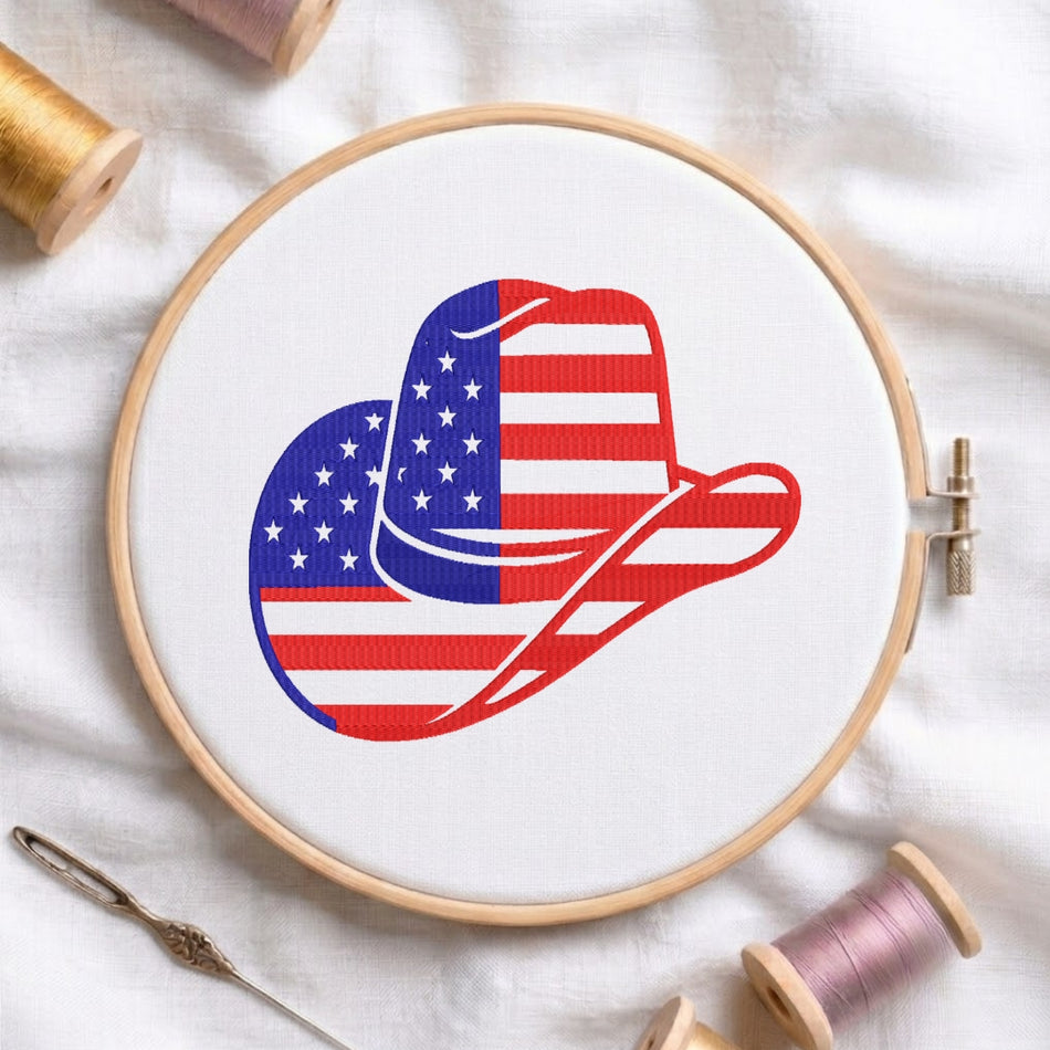 A circular embroidery hoop on white fabric, featuring a cowboy hat design filled with an American flag pattern in red, white, and blue. Spools of thread and a needle are arranged around it.