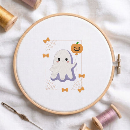  Cute ghost holding a jack-o-lantern pumpkin machine embroidery design displayed in a wooden embroidery hoop on white fabric with thread spools and a needle   