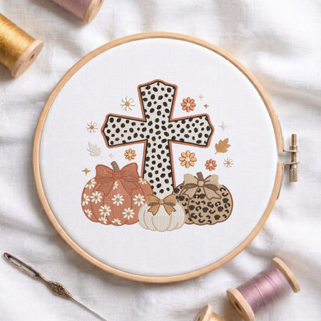 A circular embroidery hoop displays a fall-themed design with a black-and-white polka dot cross, patterned pumpkins, and small flowers on white fabric, surrounded by spools of thread and a needle.