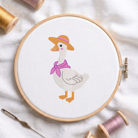  Cute duck wearing a straw hat and purple scarf machine embroidery design displayed in a wooden hoop on white fabric with thread spools and a needle   