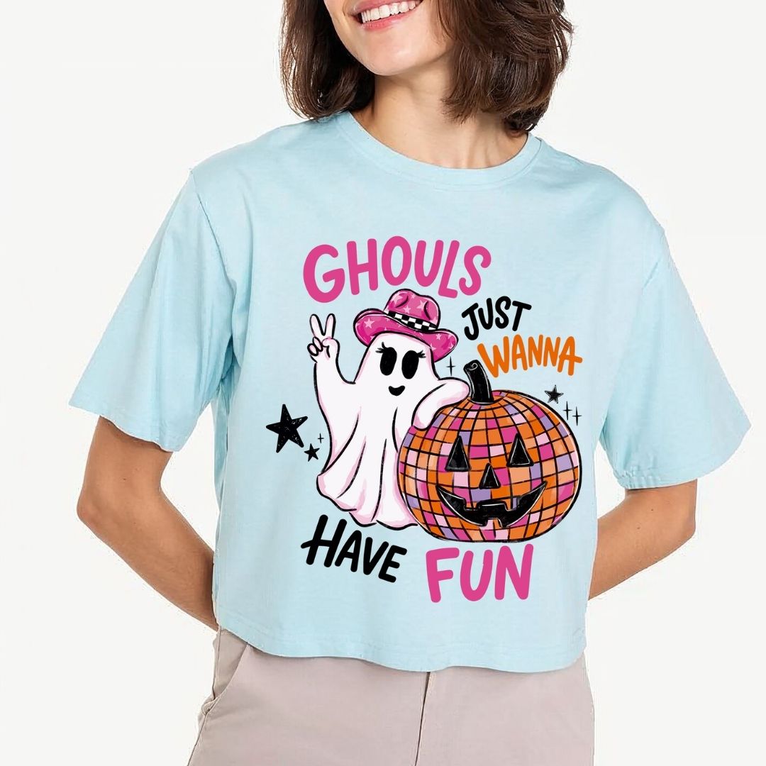 A person wearing a light blue crop top featuring an illustration of a ghost wearing a pink cowboy hat and making a peace sign next to a disco ball jack-o-lantern with the text Ghouls Just Wanna Have Fun.