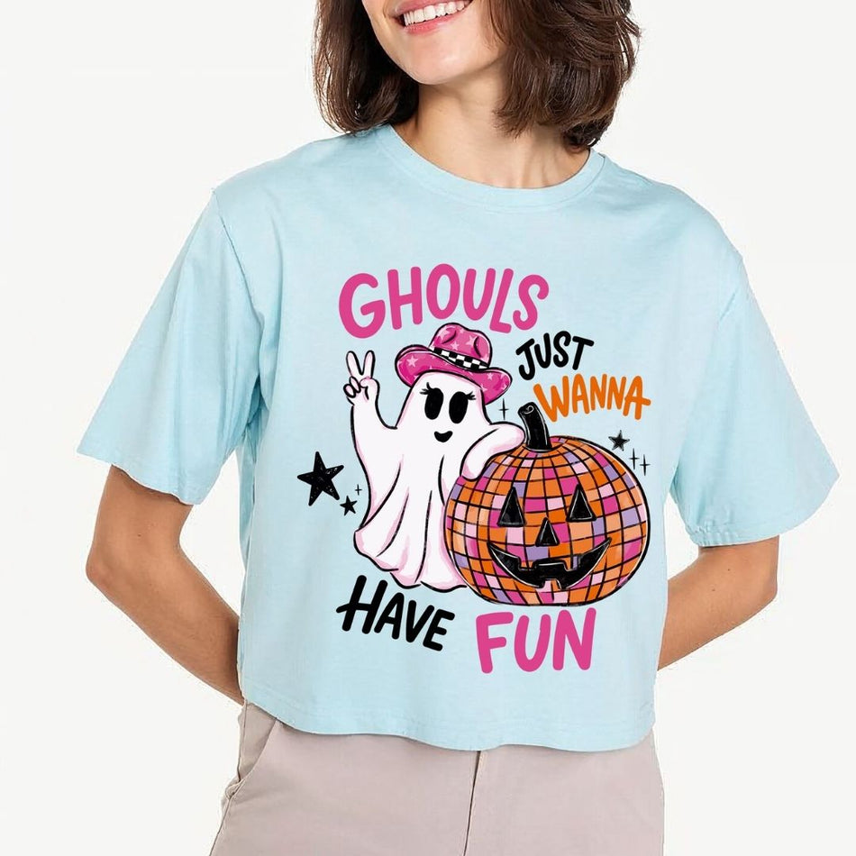 A person wearing a light blue crop top featuring an illustration of a ghost wearing a pink cowboy hat and making a peace sign next to a disco ball jack-o-lantern with the text Ghouls Just Wanna Have Fun.