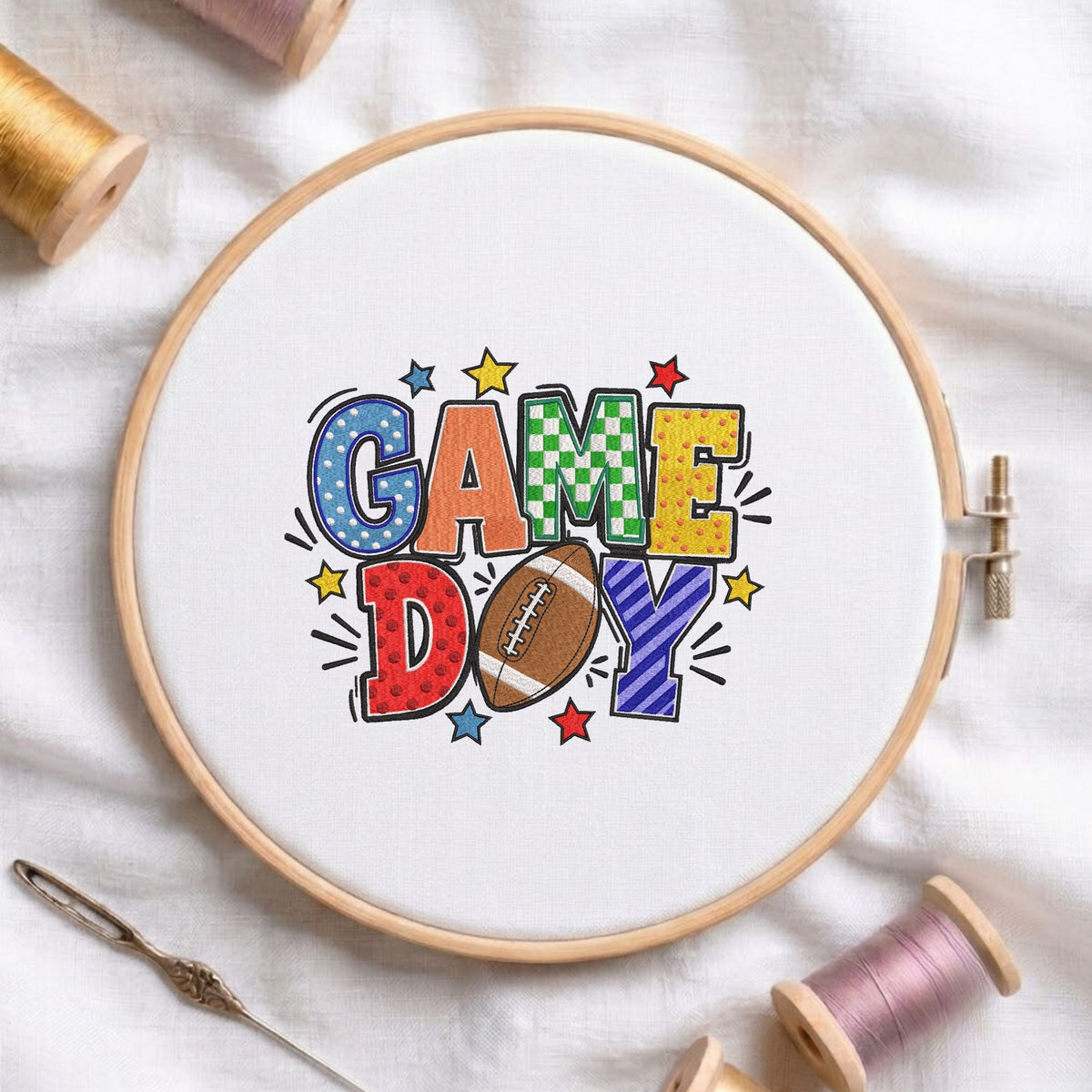 Game Day" football typography machine embroidery design displayed in a wooden hoop on white fabric with thread spools and a needle