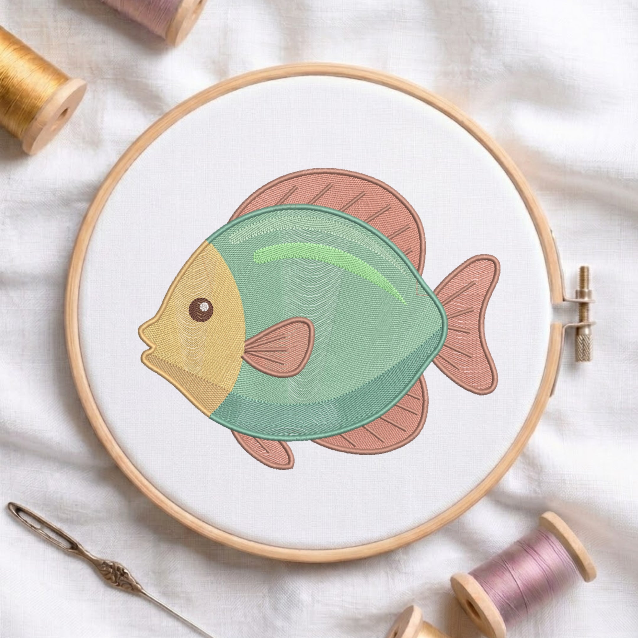 A circular embroidery hoop holds white fabric with a stylized fish design in mint green, peach, and mustard yellow. Spools of thread and a needle are arranged around it on a white fabric background.