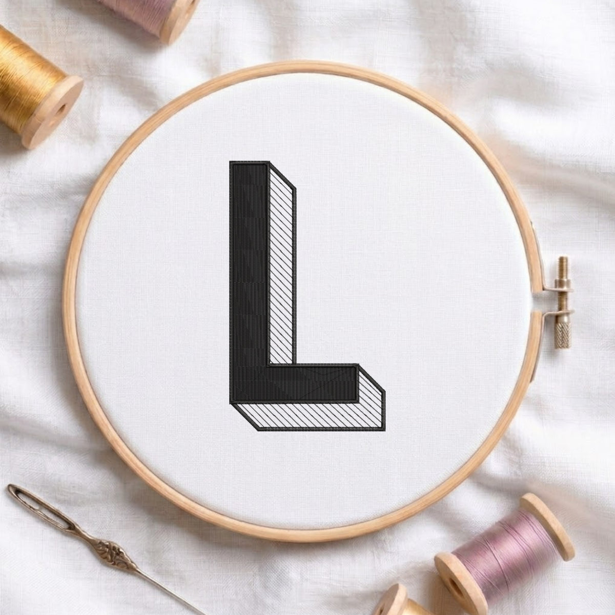 A wooden embroidery hoop holding white fabric with a black embroidered letter L, surrounded by spools of thread and a needle on a white fabric background.