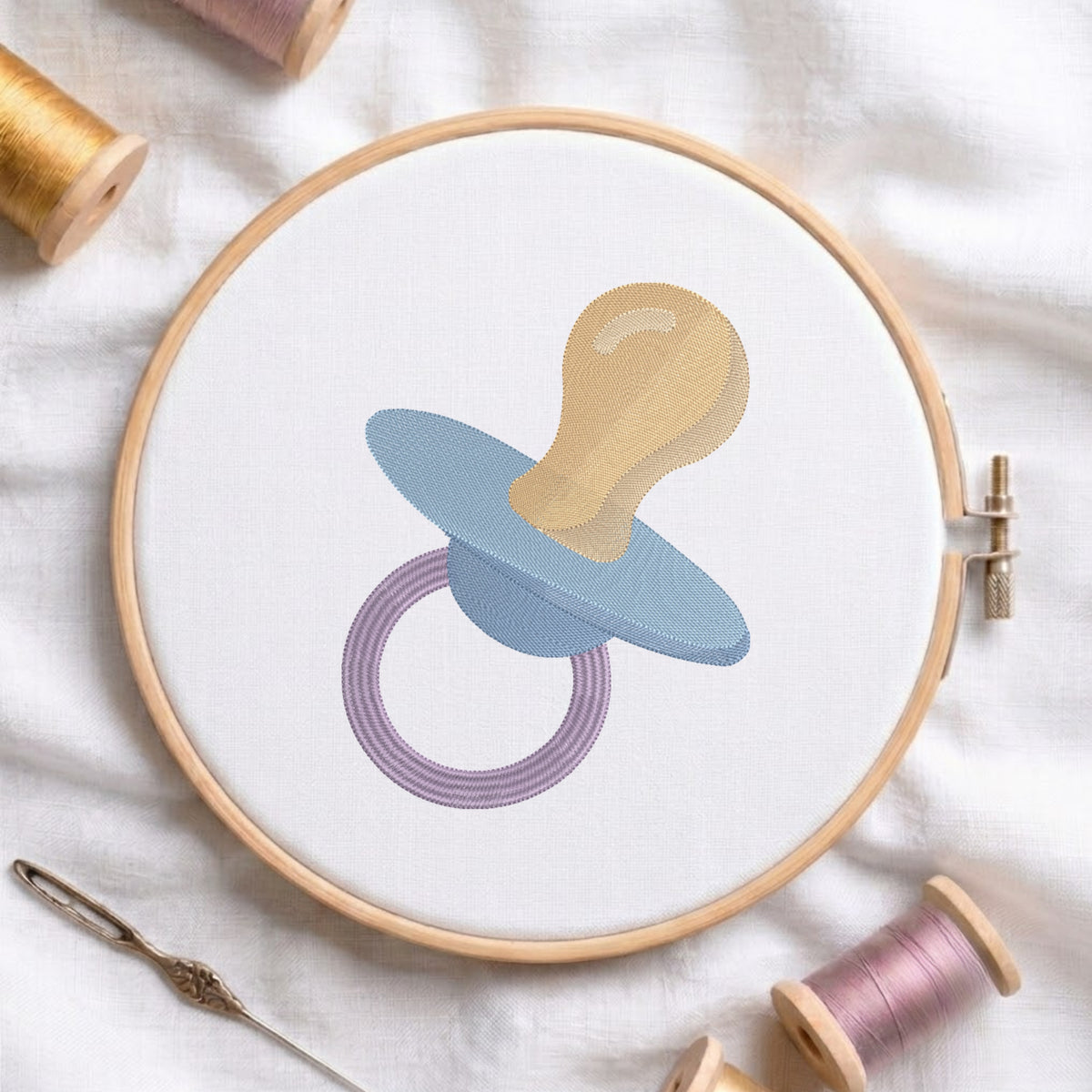 A circular embroidery hoop holding white fabric with a stylized pacifier design in light blue, beige, and lavender. The hoop is styled on a white fabric background with embroidery threads, a needle, and a thread spool nearby.