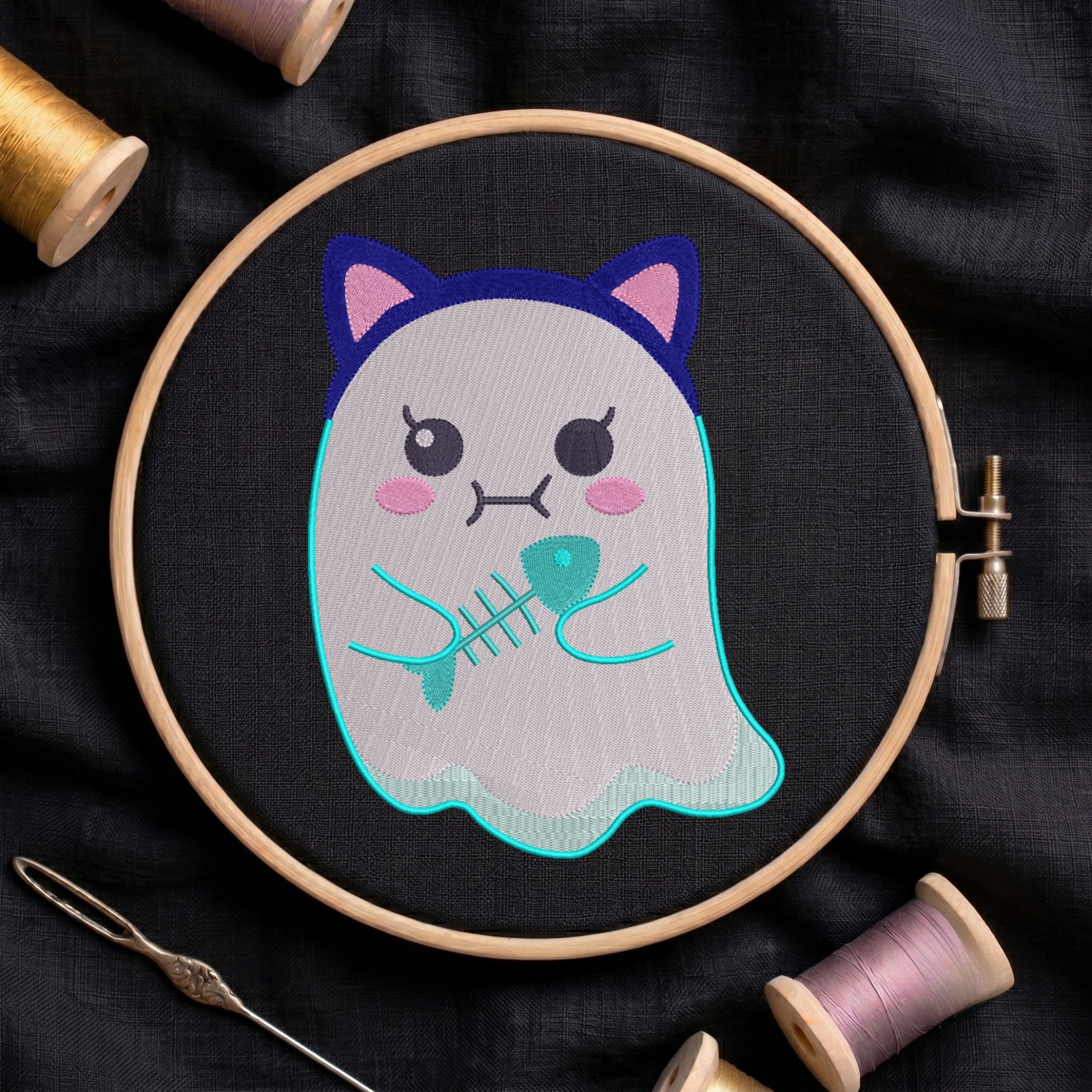 A close-up of a cute ghost wearing cat ears and holding a fish skeleton embroidery design, stitched on black fabric inside a wooden embroidery hoop surrounded by spools of thread and a needle.