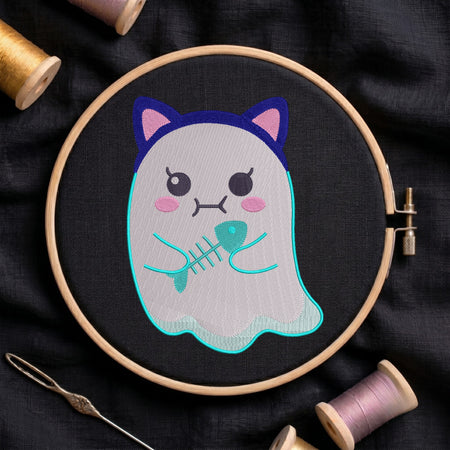 A close-up of a cute ghost wearing cat ears and holding a fish skeleton embroidery design, stitched on black fabric inside a wooden embroidery hoop surrounded by spools of thread and a needle.