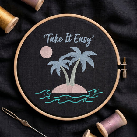 A circular embroidery hoop holding a black fabric piece with a stitched tropical island design and the words “Take It Easy” in light blue script. The design includes two palm trees, a pink sun, a pink island, and turquoise waves.