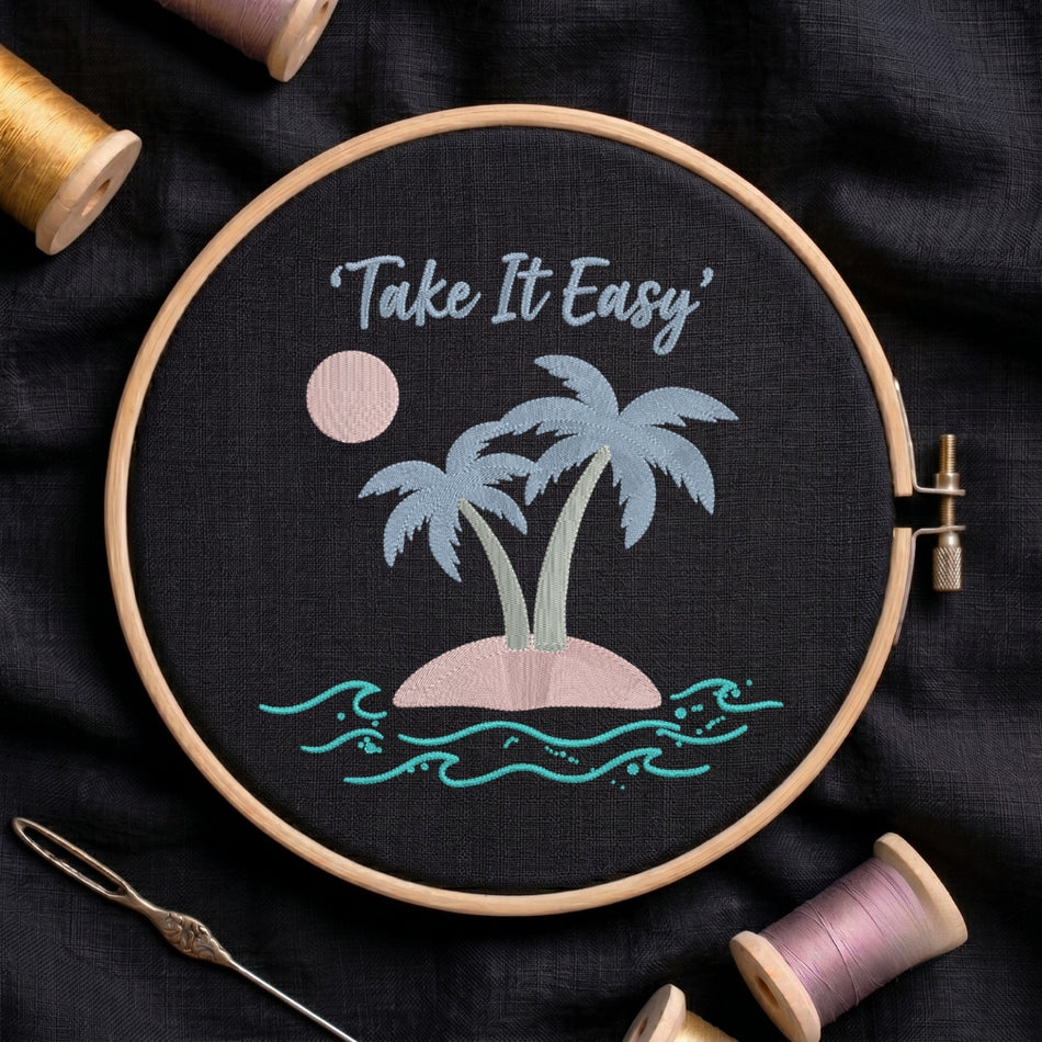 A circular embroidery hoop holding a black fabric piece with a stitched tropical island design and the words “Take It Easy” in light blue script. The design includes two palm trees, a pink sun, a pink island, and turquoise waves.