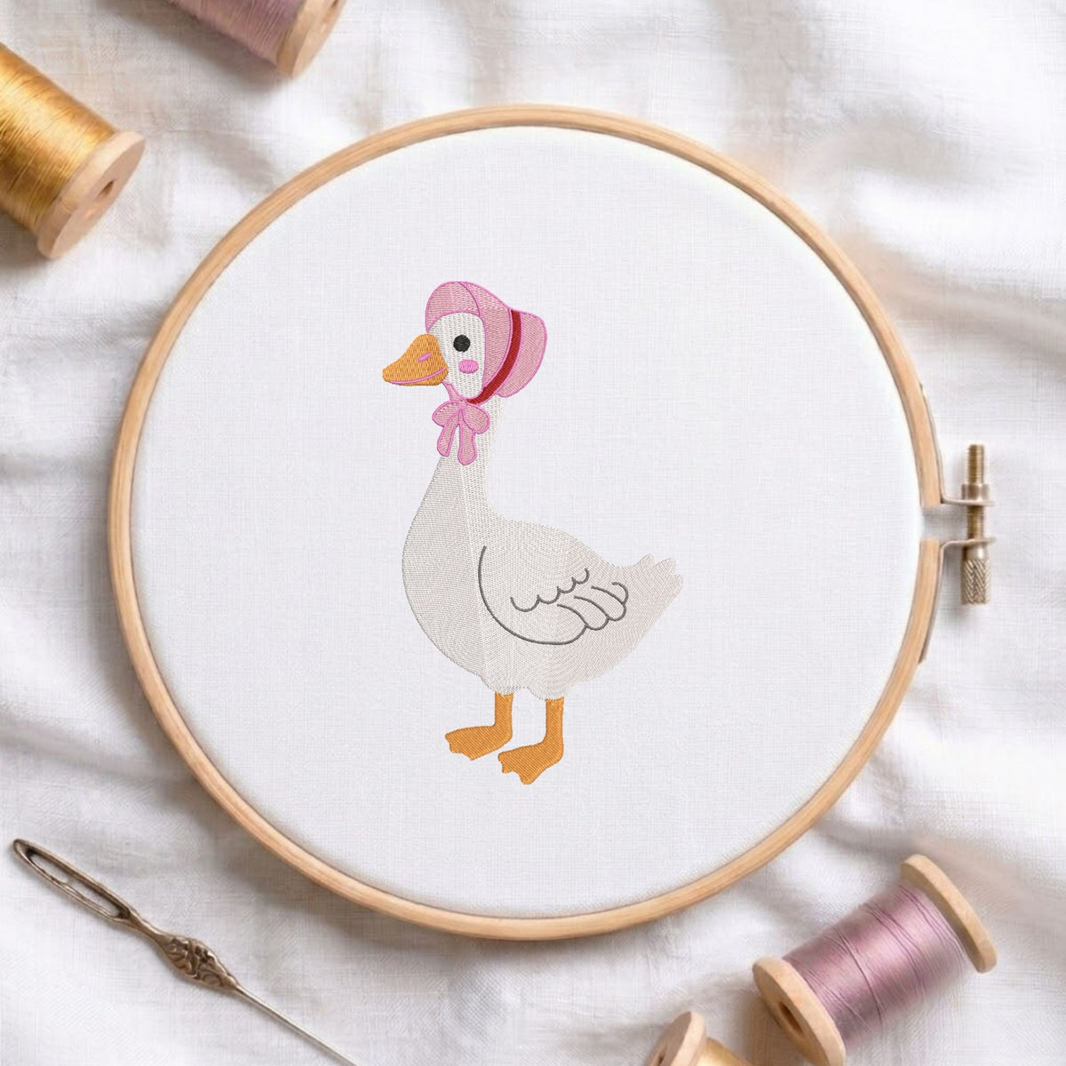 Cute goose wearing a pink bonnet machine embroidery design displayed in a wooden hoop on white fabric with thread spools and a needle.