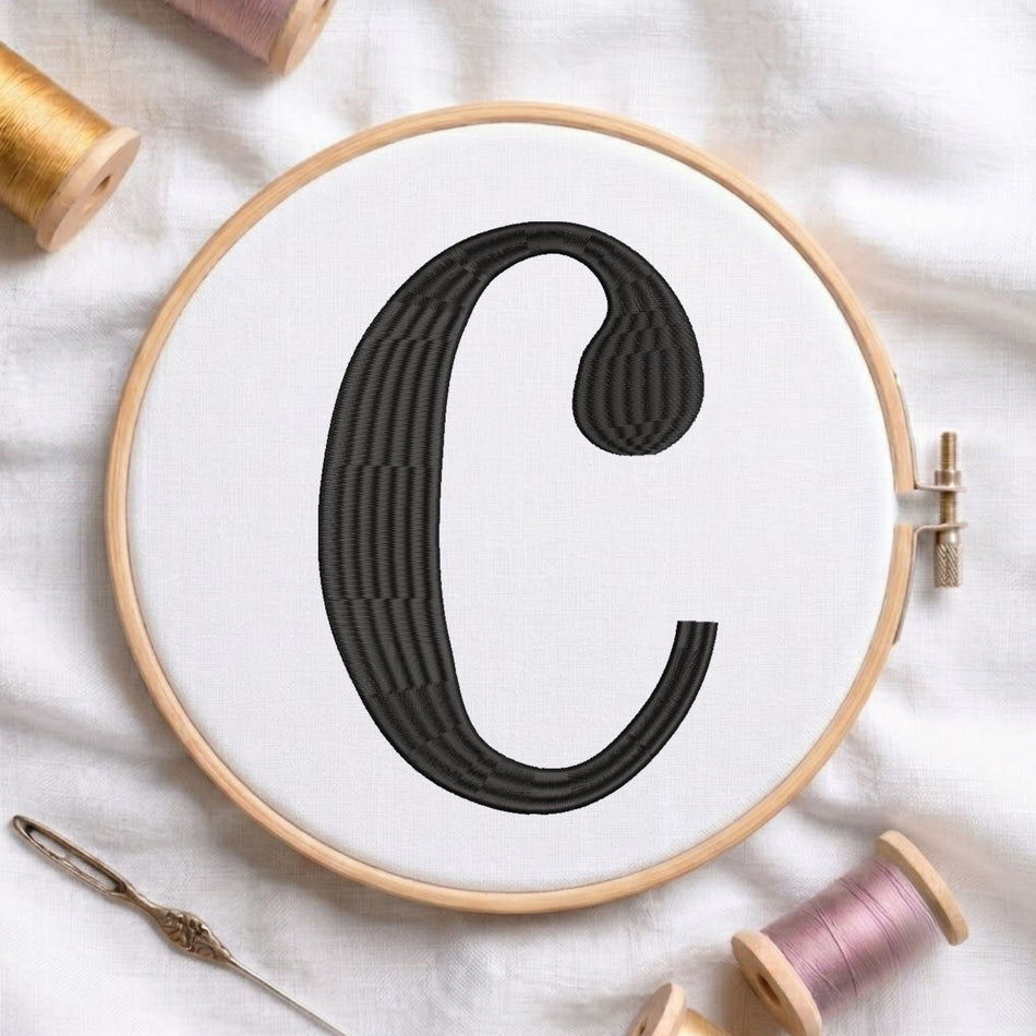 A wooden embroidery hoop holding white fabric with a large black embroidered letter C, styled on a white fabric background with spools of thread and a needle.