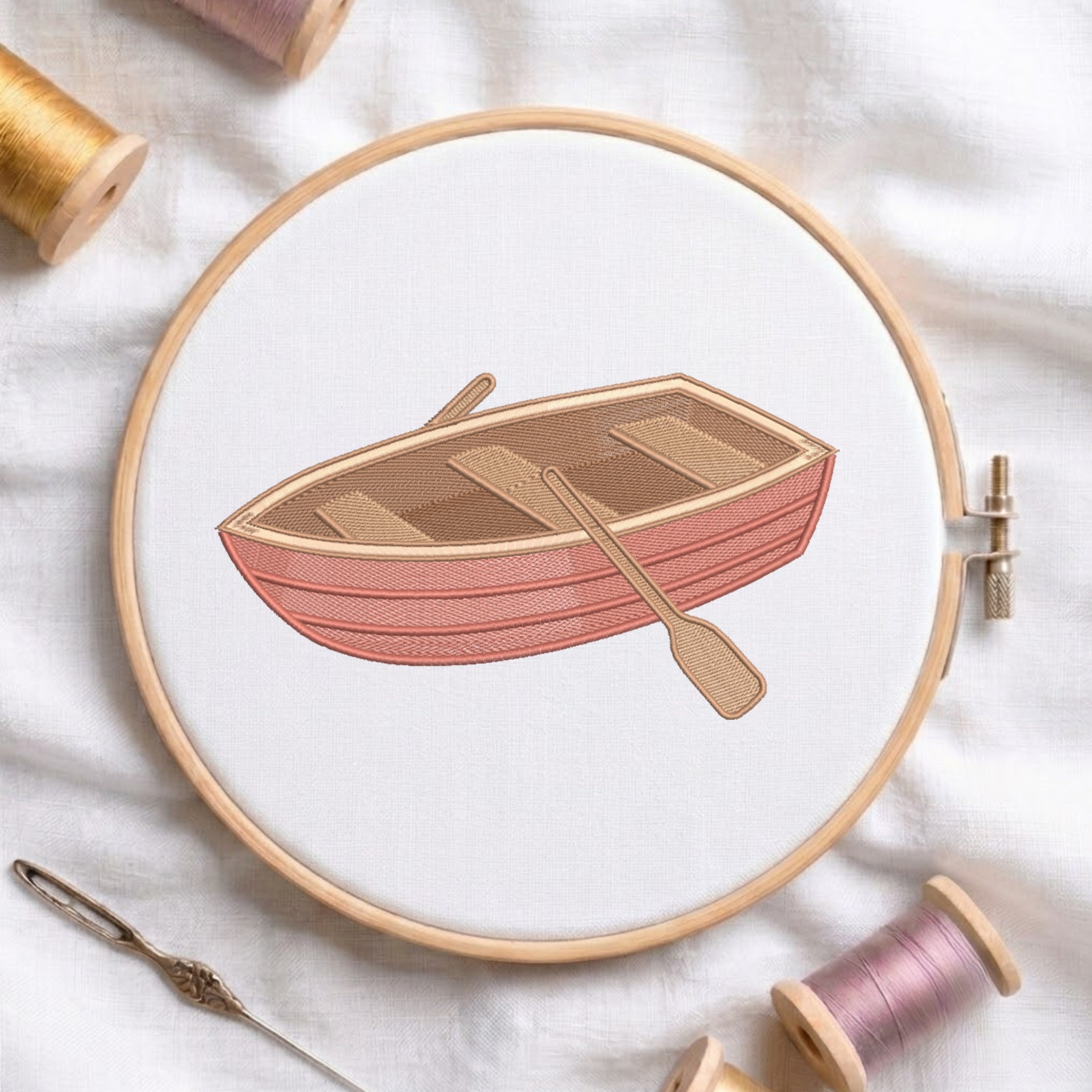 A circular embroidery hoop holds a white fabric with a stitched illustration of a pink and tan rowboat and oar. Spools of thread, a needle, and soft fabric are arranged around it in a styled flat-lay.