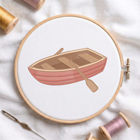 A circular embroidery hoop holds a white fabric with a stitched illustration of a pink and tan rowboat and oar. Spools of thread, a needle, and soft fabric are arranged around it in a styled flat-lay.