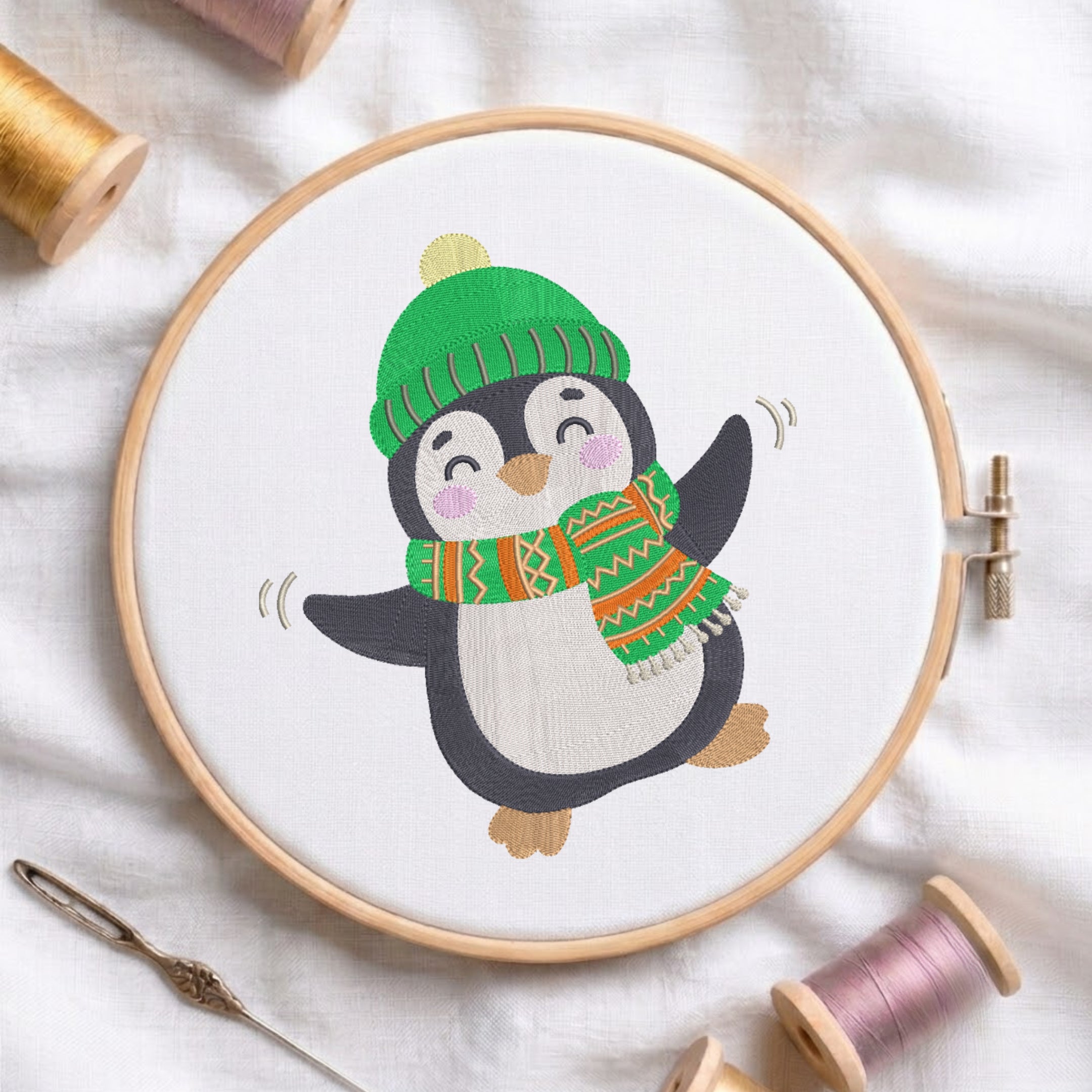 A circular embroidery hoop holds a white fabric with a cartoon penguin design wearing a green striped beanie and a multicolor patterned scarf. The hoop is photographed on white fabric with spools of thread and a needle around it.