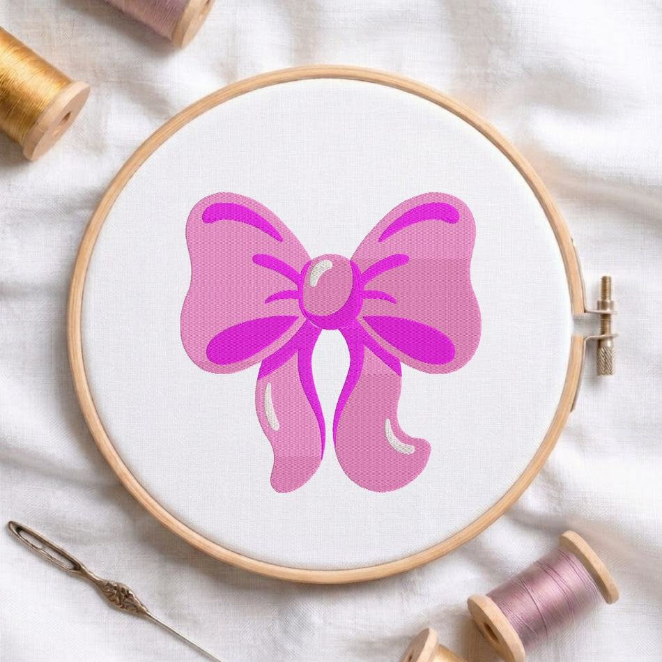 A pink bow design displayed inside a wooden embroidery hoop on white fabric, surrounded by spools of thread and a needle on a soft white background.