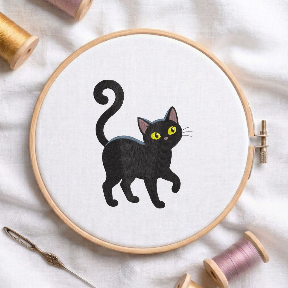 Cute walking black cat with big yellow eyes and a curved tail machine embroidery design, displayed inside a round wooden hoop on white fabric with thread spools and a needle.