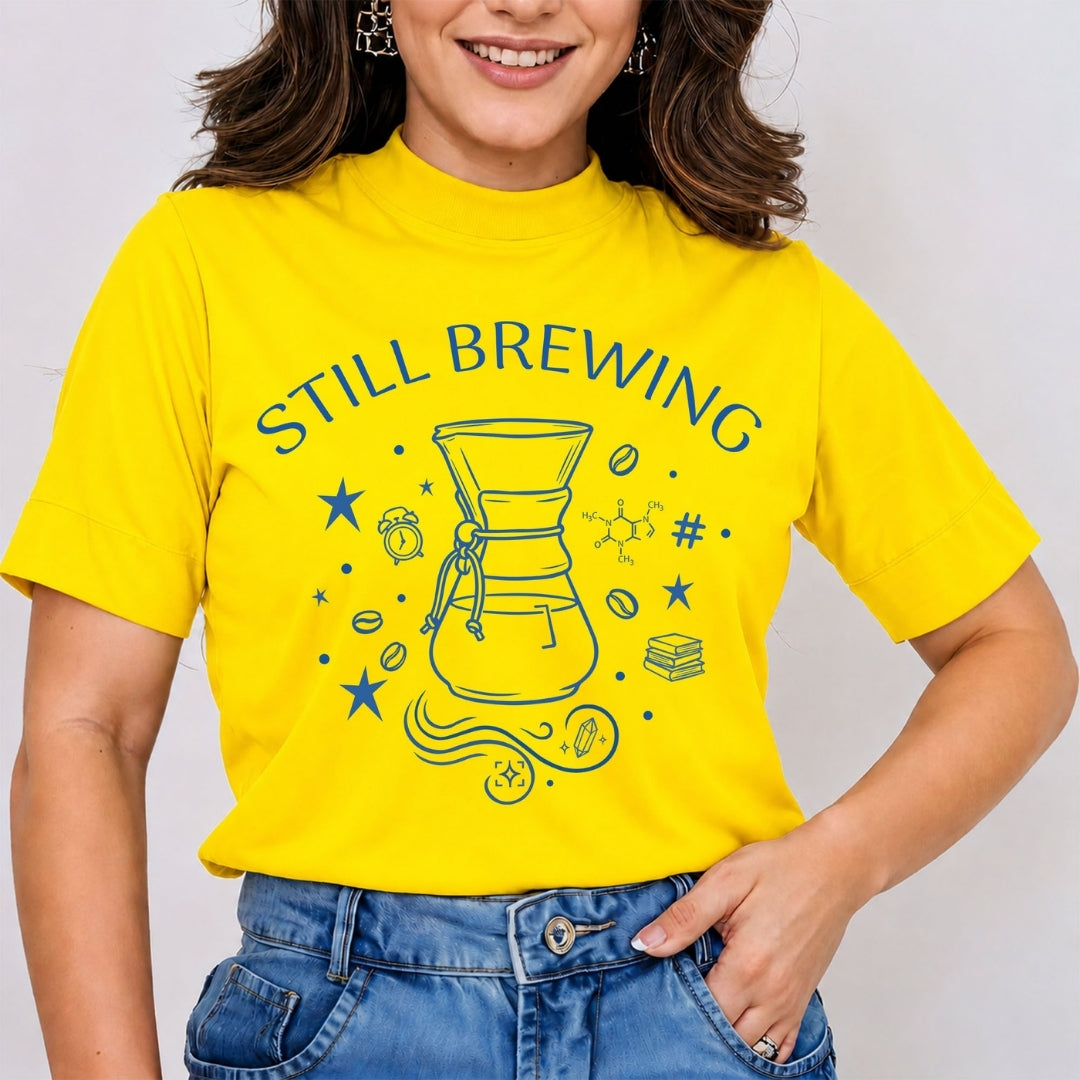 Woman wearing a bright yellow mock neck t-shirt featuring a blue line art "Still Brewing" graphic print with a pour-over coffee carafe, stars, and coffee beans, tucked into blue jeans.