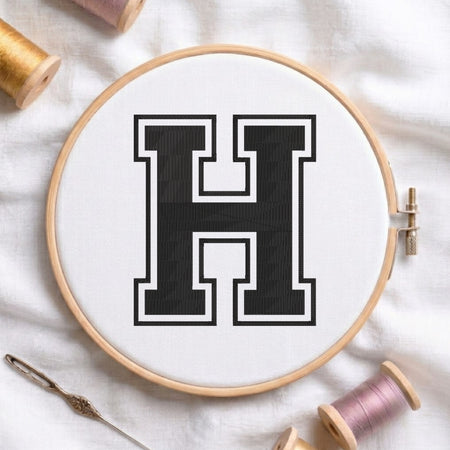 A wooden embroidery hoop holding white fabric with a large black letter H outlined in white, styled on a white cloth with spools of thread and a needle nearby.