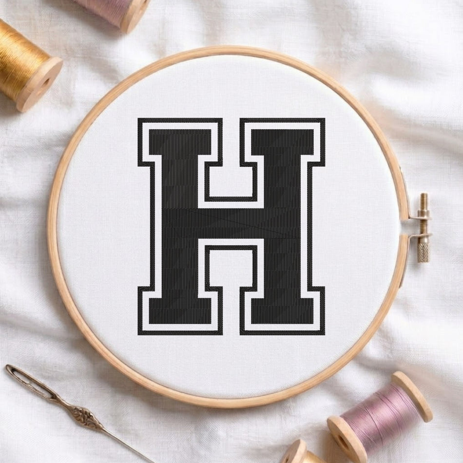 A wooden embroidery hoop holding white fabric with a large black letter H outlined in white, styled on a white cloth with spools of thread and a needle nearby.