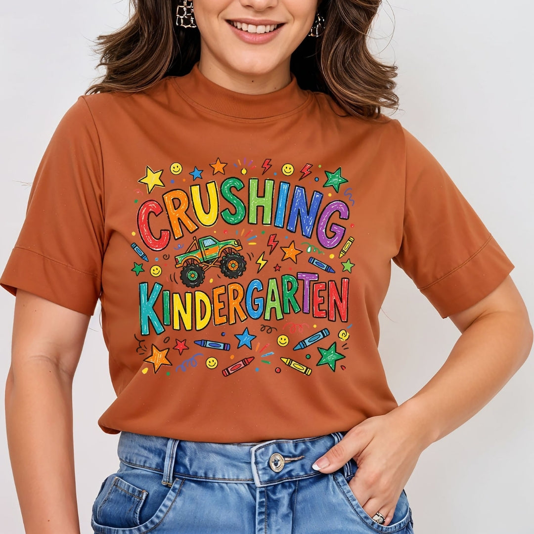 A woman wearing a rust-orange short-sleeve t-shirt with a colorful graphic that reads 'CRUSHING KINDERGARTEN' in multicolored letters, featuring a green monster truck and school-themed doodles, paired with blue jeans.