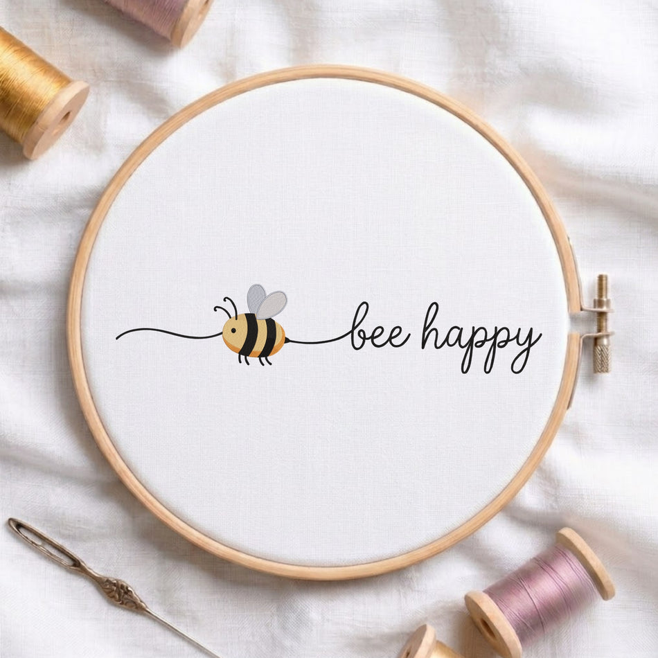 Bee happy" quote with a cute flying bee machine embroidery design displayed inside a round wooden hoop on white linen fabric, accompanied by wooden thread spools and a needle.