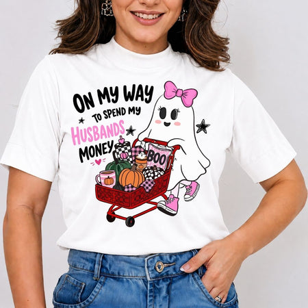 A person wearing a white t-shirt featuring a cute ghost pushing a red shopping cart filled with pumpkins, a coffee cup, and a "BOO" pillow, with the text "ON MY WAY TO SPEND MY HUSBANDS MONEY".
