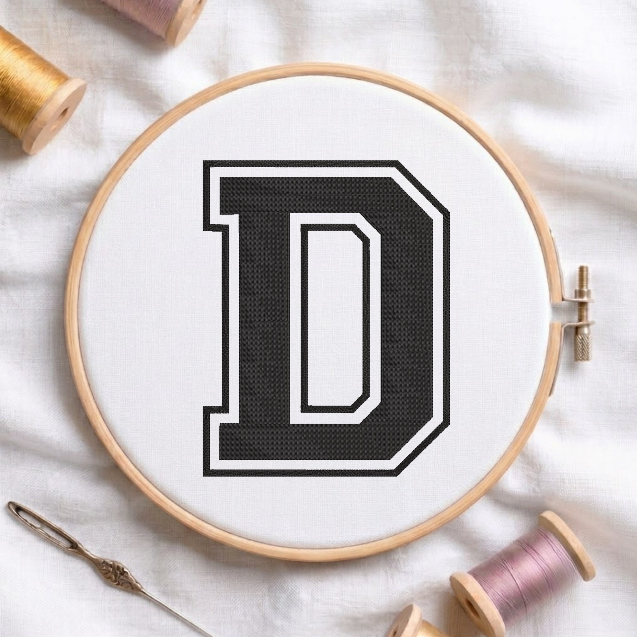 A wooden embroidery hoop holding white fabric with a large black letter D outlined in white, styled on a white cloth with thread spools and a needle around it.