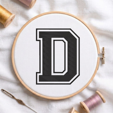 A wooden embroidery hoop holding white fabric with a large black letter D outlined in white, styled on a white cloth with thread spools and a needle around it.