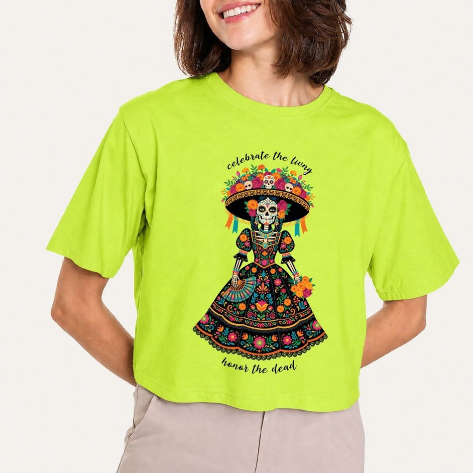 A woman wearing a bright lime green cropped t-shirt with a colorful Day of the Dead (Dia de los Muertos) sugar skull graphic on the front, paired with light beige pants, against a plain light background.